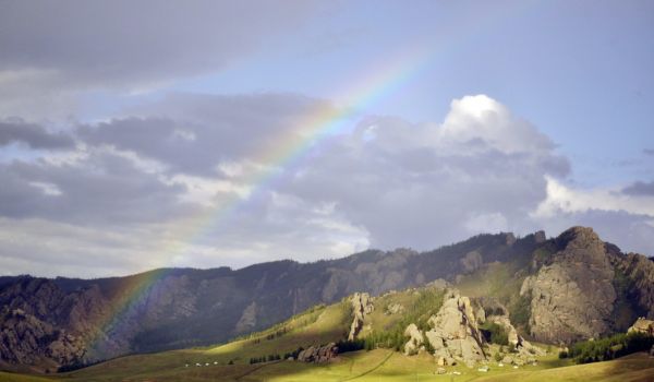 Landschaft mit hohen Bergen und grünen Wiesen. Im Hintergrund leichte Wolken und ein Regenbogen.